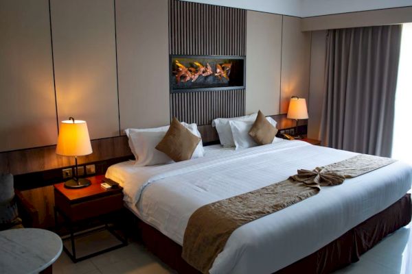 The image shows a modern hotel room with a king-size bed, two bedside tables with lamps, a wall-mounted fireplace, and curtains covering a window.