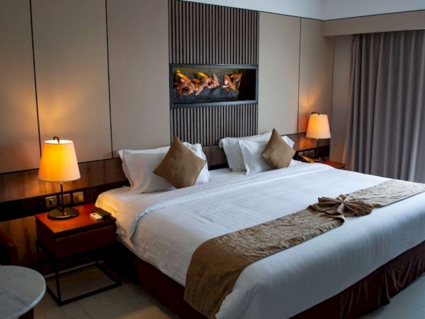 The image shows a modern hotel room with a king-size bed, two bedside tables with lamps, a wall-mounted fireplace, and curtains covering a window.