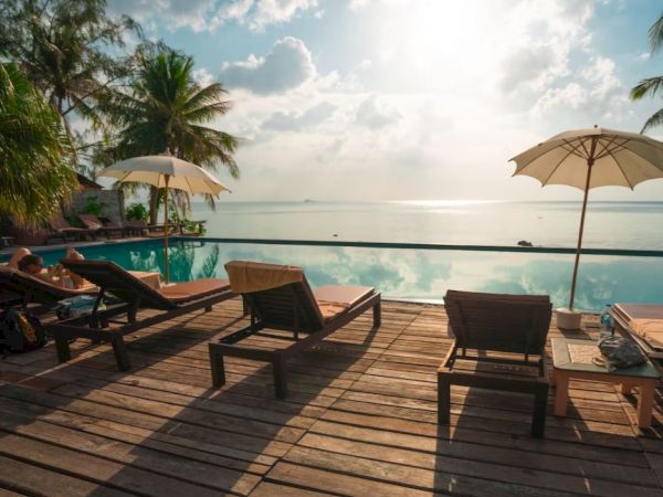 Sun loungers and umbrellas on a wooden deck overlook an infinity pool and an ocean view, with palm trees and a serene sky in the background.
