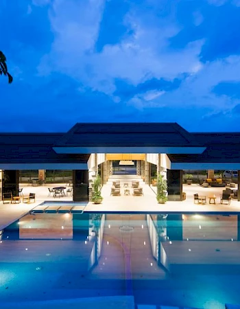 A luxurious, illuminated outdoor pool area at night with a modern building in the background, featuring seating and dining areas.
