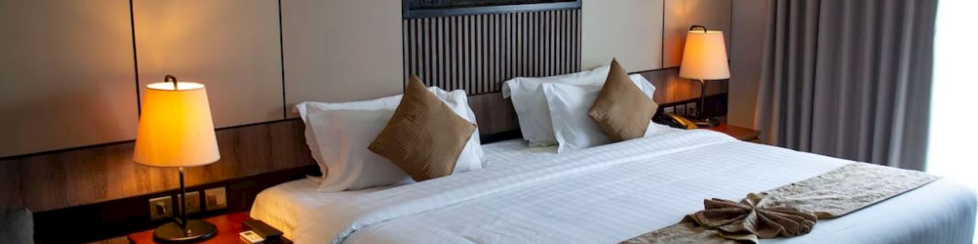 A modern hotel room with a large bed, bedside tables, and two lamps. The room is well-lit and has a decorative fireplace.