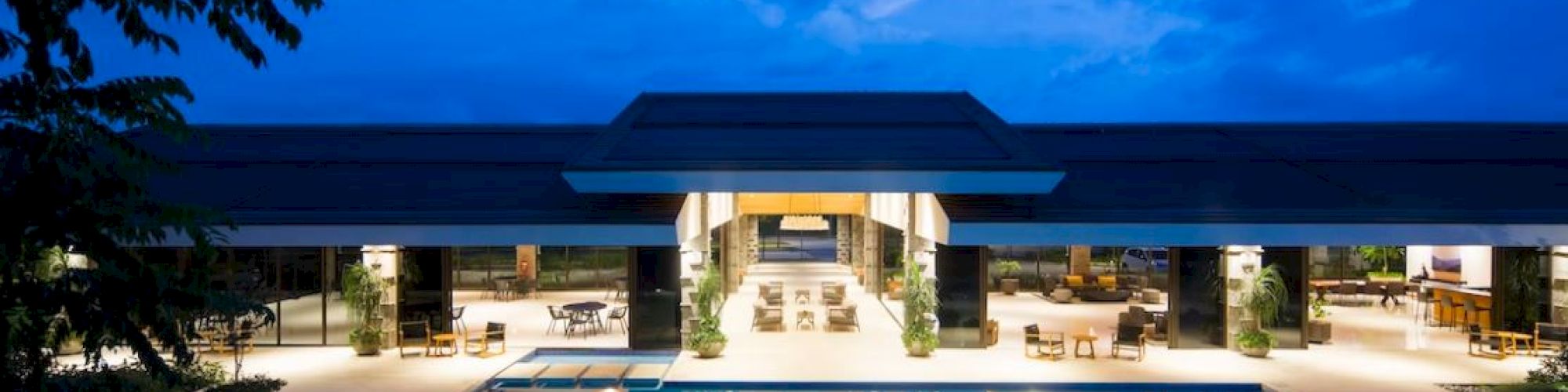 The image shows a luxurious, illuminated pool area with a modern building in the background, surrounded by seating and trees under a twilight sky.