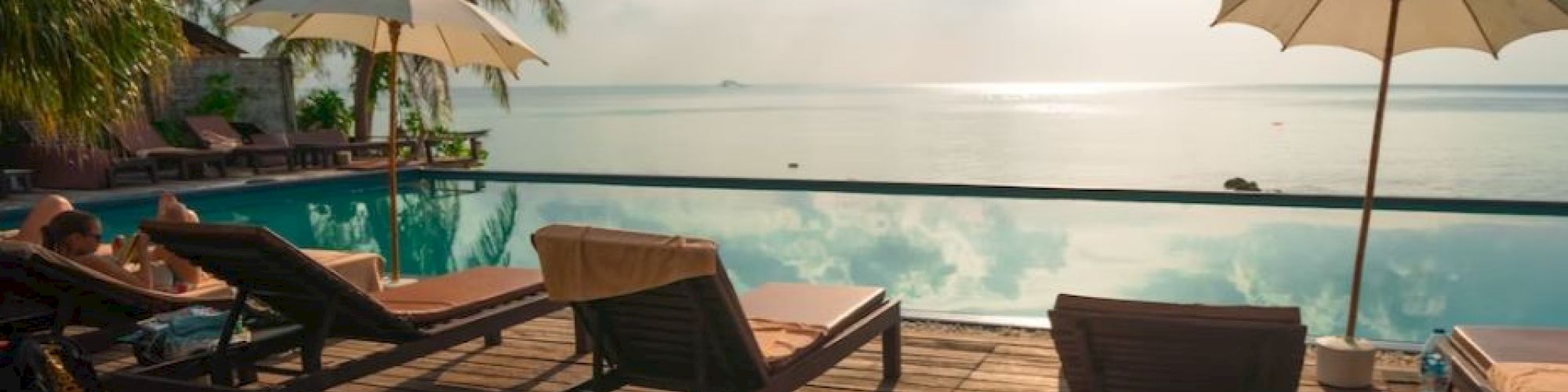 A serene lounge area with sunbeds and umbrellas by a pool, overlooking a calm sea under a partly cloudy sky and bright sunlight.