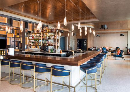 This image shows a modern bar with a well-stocked liquor shelf, blue bar stools, and stylish pendant lights in a comfortable and spacious setting.