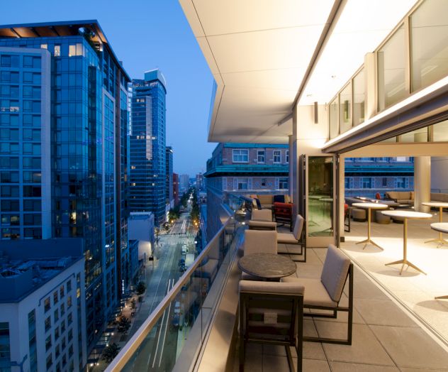 The image shows a modern urban balcony at dusk, overlooking a city street with tall buildings, furnished with tables and chairs.