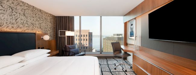 A modern hotel room with a large bed, desk, chair, flat-screen TV, and a window offering a city view. The room has a stylish and clean design.