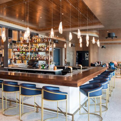 This image shows a modern bar with a wooden counter, blue barstools, pendant lights, and a well-stocked shelf of bottles behind the bar.