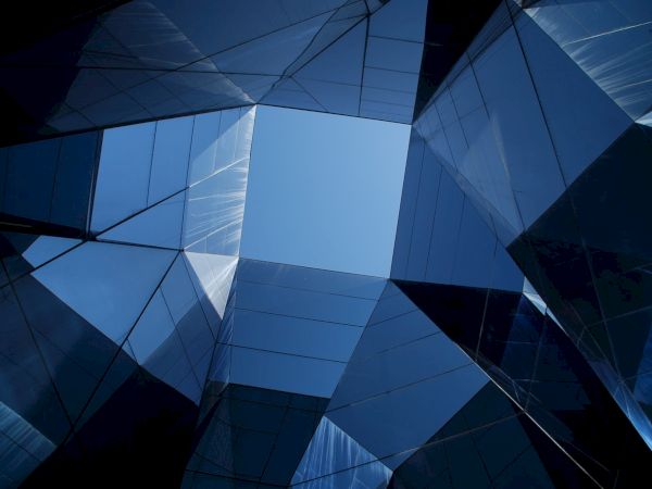The image shows a view looking up at modern reflective glass buildings forming geometric shapes, with a background of clear blue sky.
