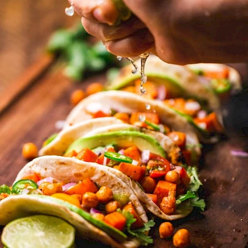 A hand is squeezing lime over a row of tacos filled with chickpeas, vegetables, and garnished with avocado slices, lined up on a wooden board.