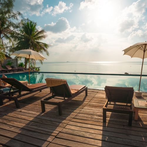 The image shows a serene poolside scene with lounge chairs, umbrellas, palm trees, and a stunning view of the ocean under a bright sky.