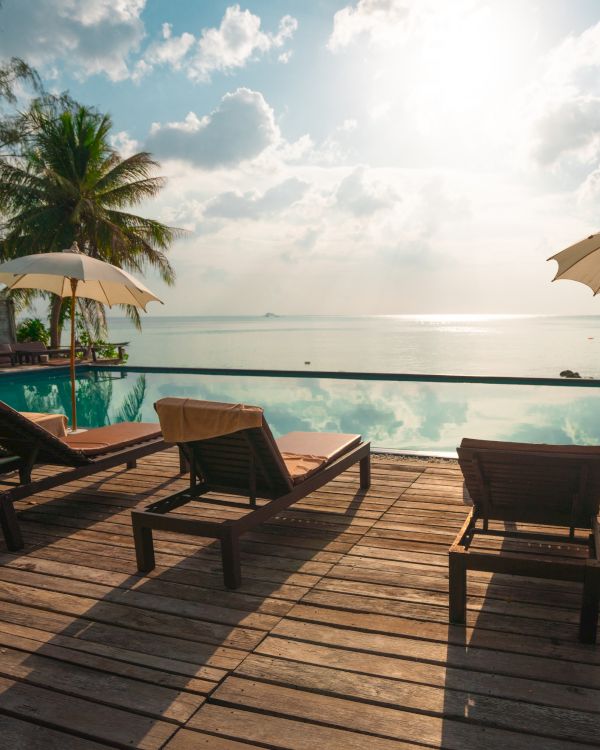 The image shows a serene poolside scene with lounge chairs, umbrellas, palm trees, and a stunning view of the ocean under a bright sky.