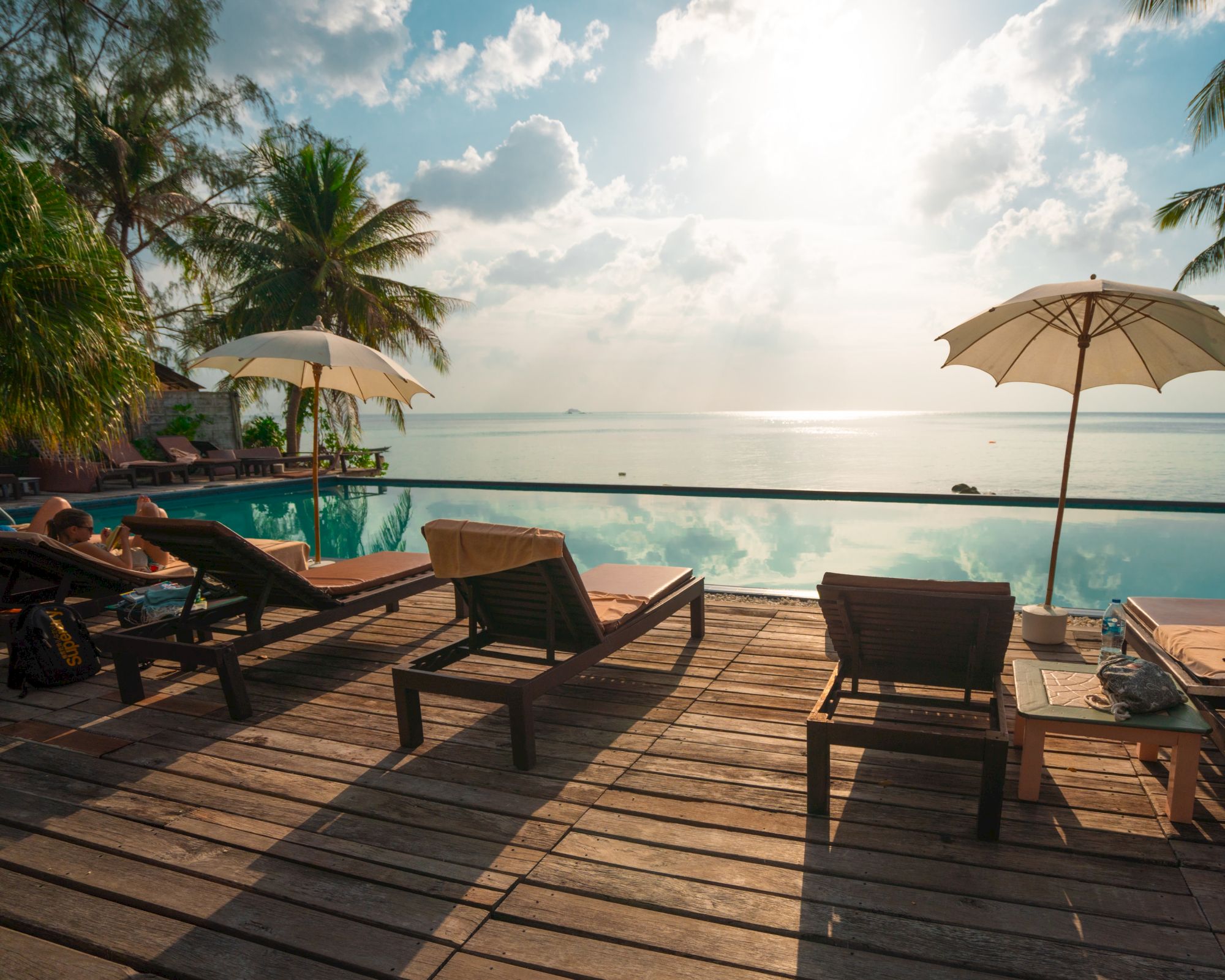 A serene tropical scene with lounge chairs and umbrellas on a wooden deck by a pool overlooking the ocean, surrounded by palm trees.