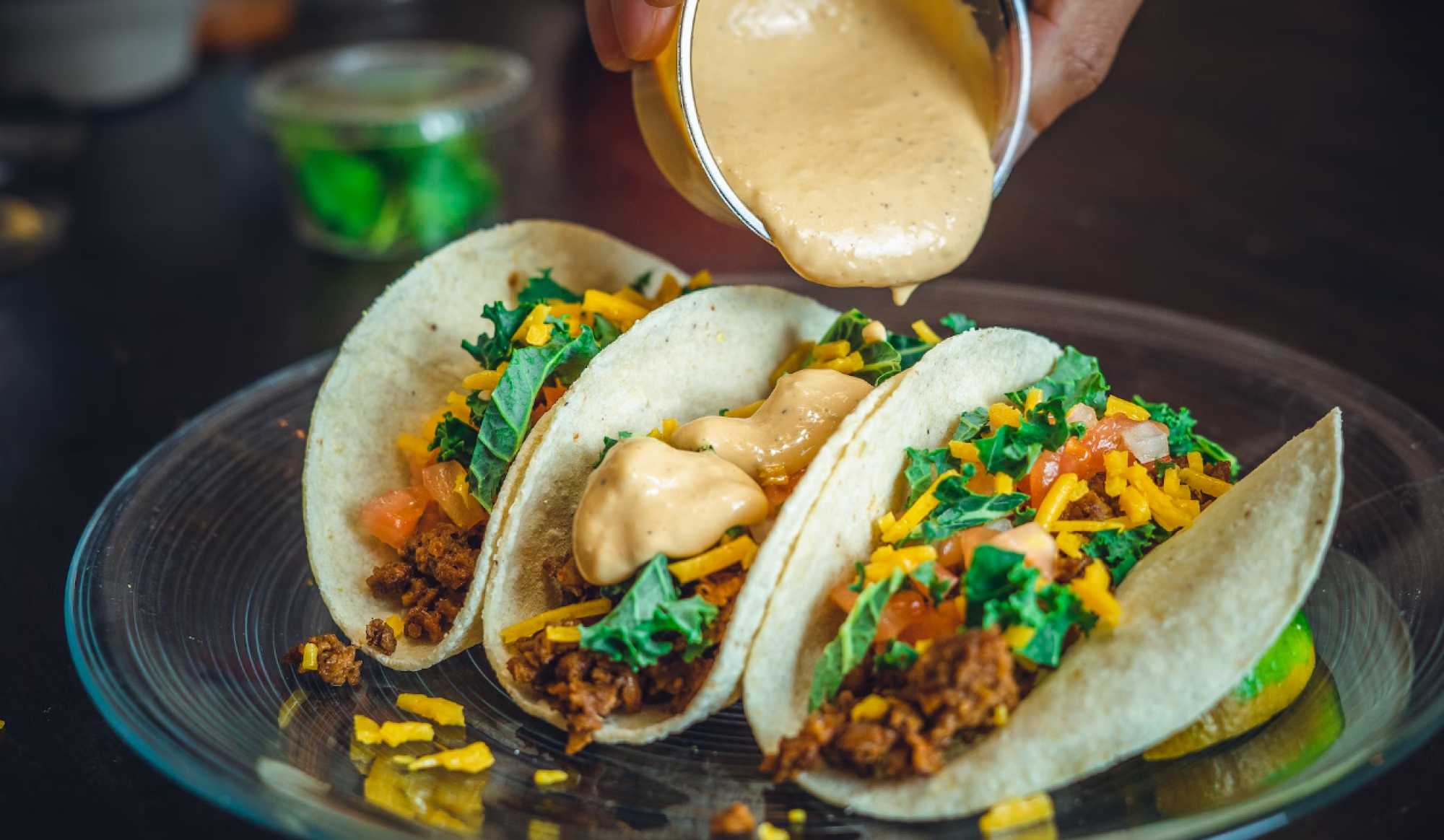 The image shows three tacos on a plate being topped with a creamy sauce. The tacos are filled with vegetables, meat, and cheese.