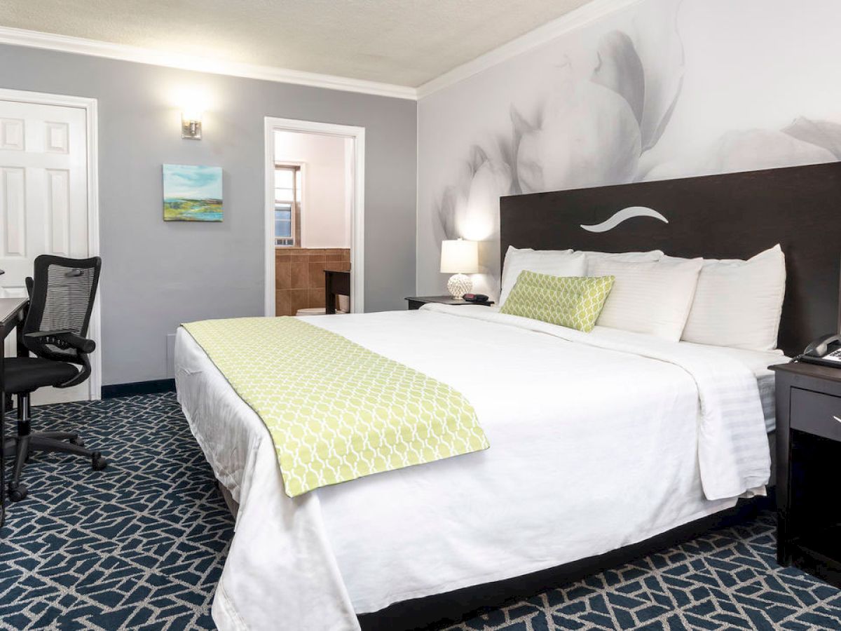 This image displays a hotel room with a king-sized bed, a work desk, two nightstands with lamps, and a modern design.