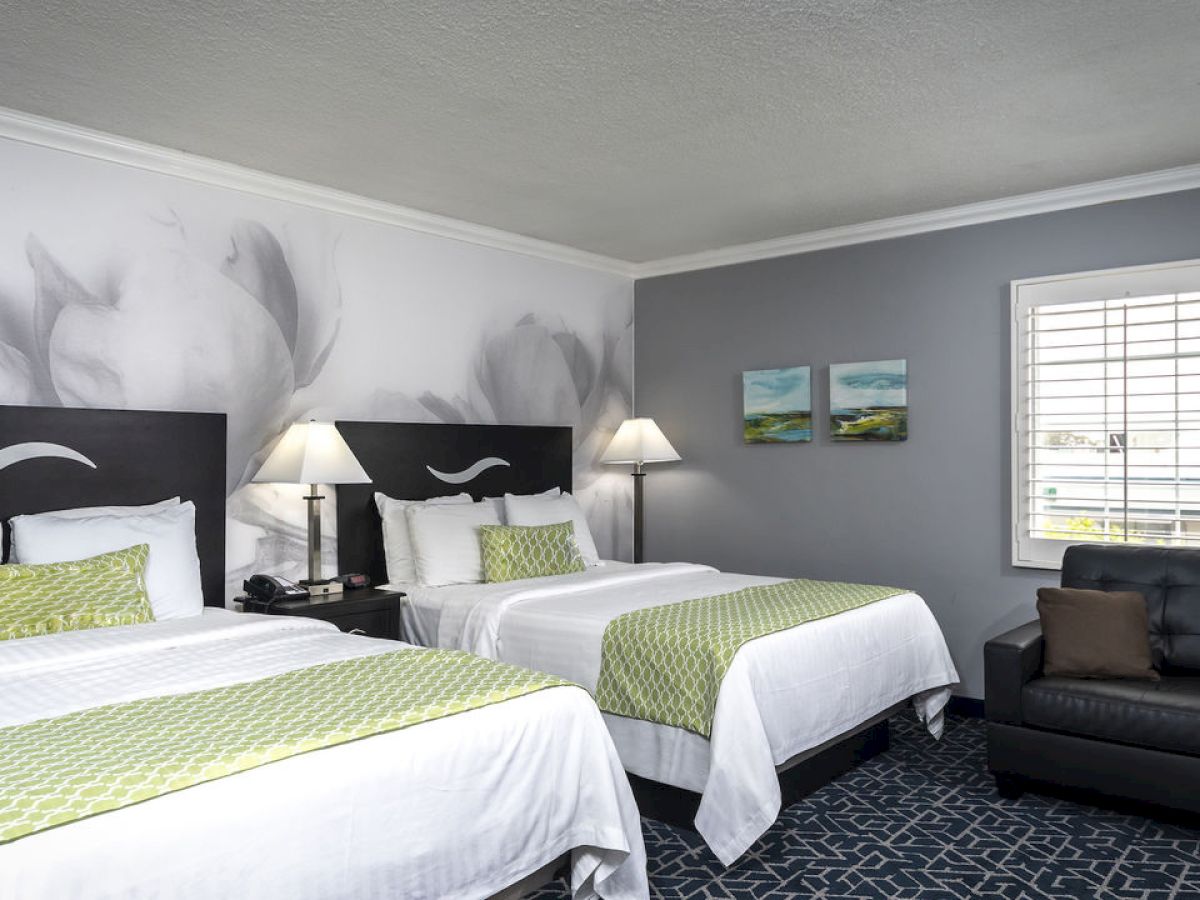 A hotel room with two beds, green and white bedding, wall art, lamps, a window with blinds, and a black sofa.