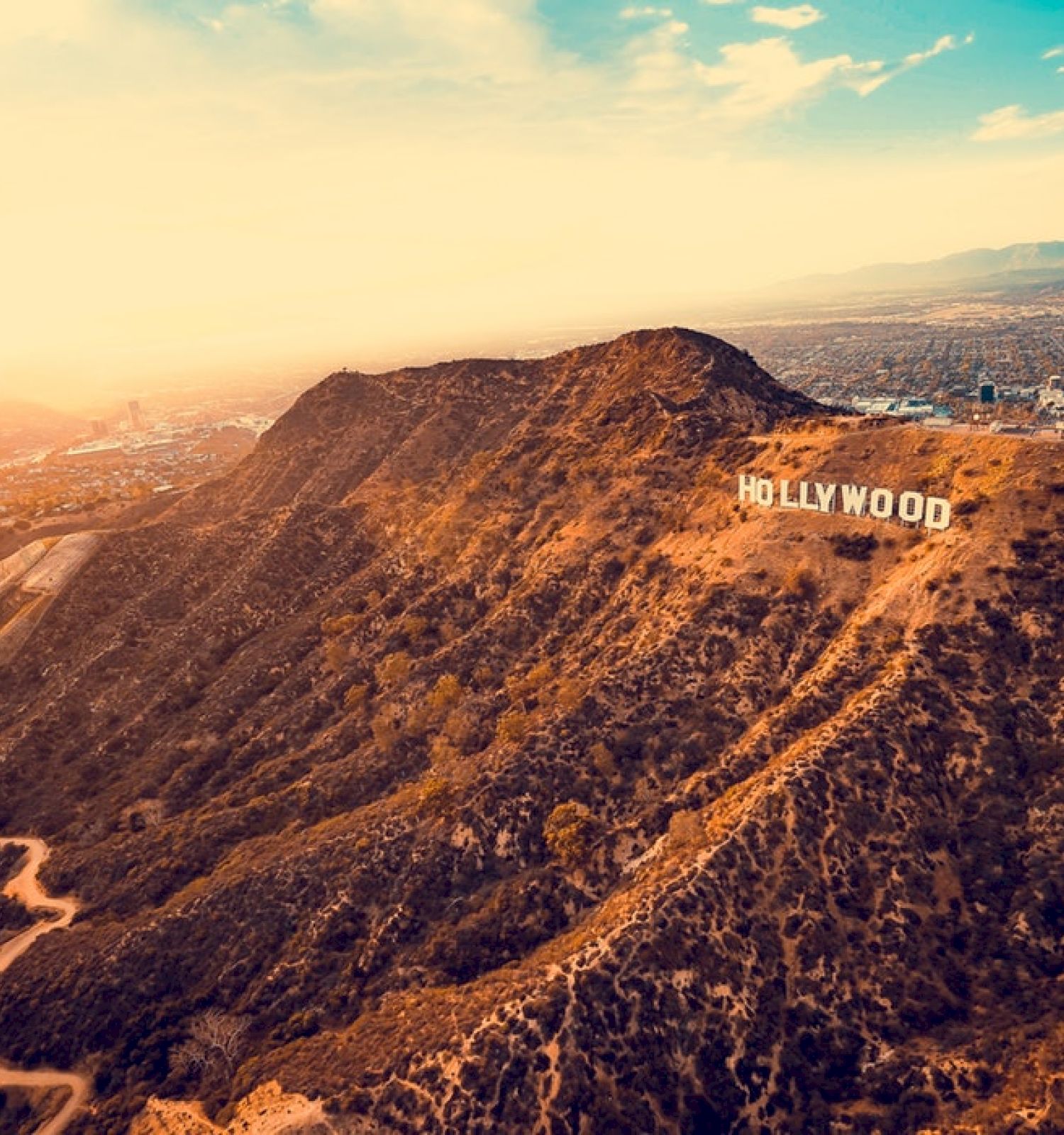 An aerial view of the Hollywood Hills with the iconic Hollywood sign displayed prominently, overlooking the city of Los Angeles during sunset.