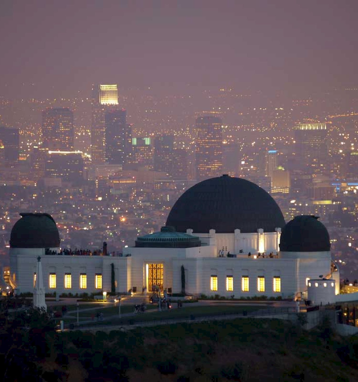 This image shows an observatory building with a large domed roof in the foreground and a cityscape with numerous illuminated high-rise buildings behind it.