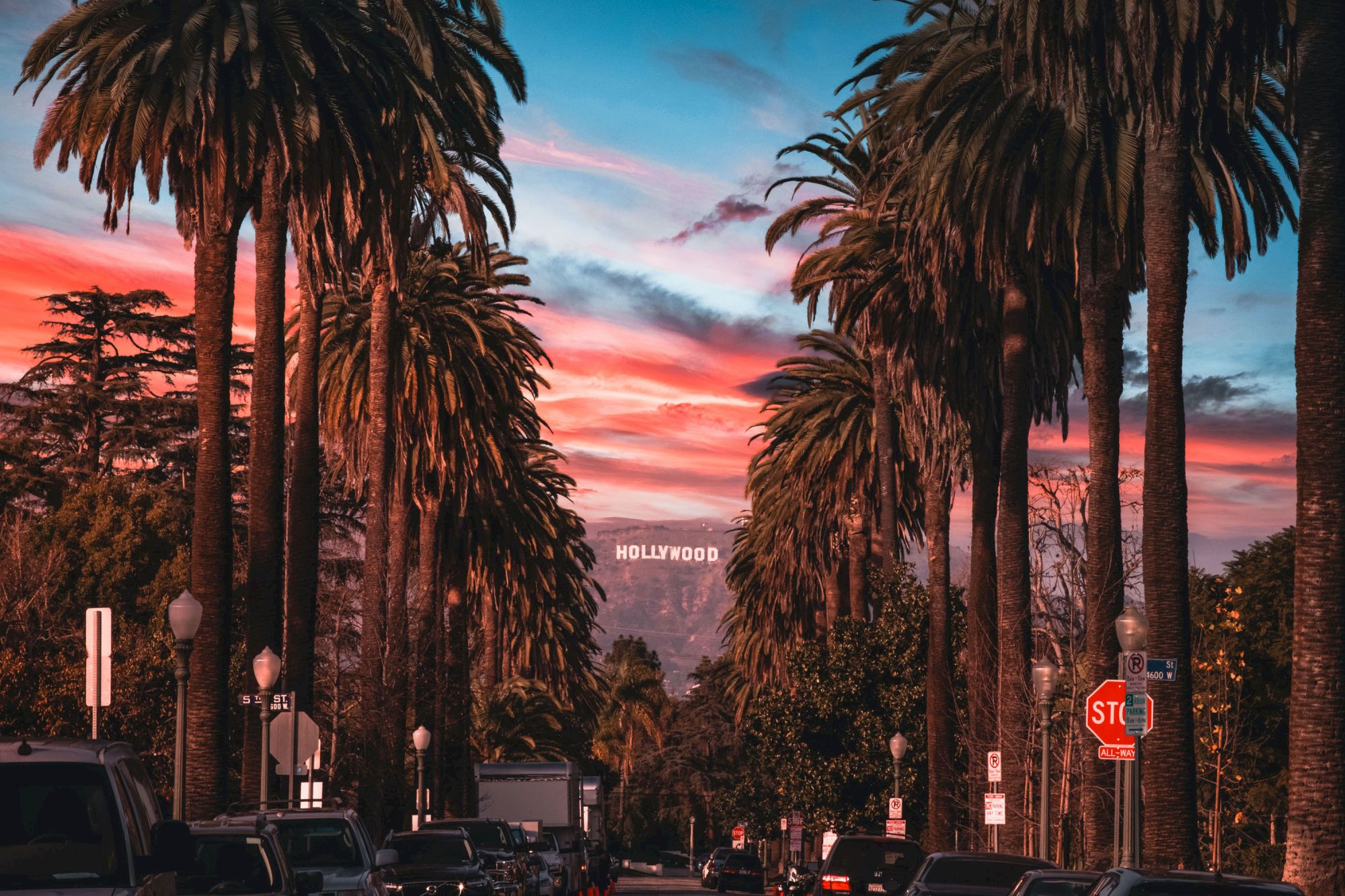 A street lined with tall palm trees and parked cars, with the iconic Hollywood sign visible in the distance against a colorful sunset sky.