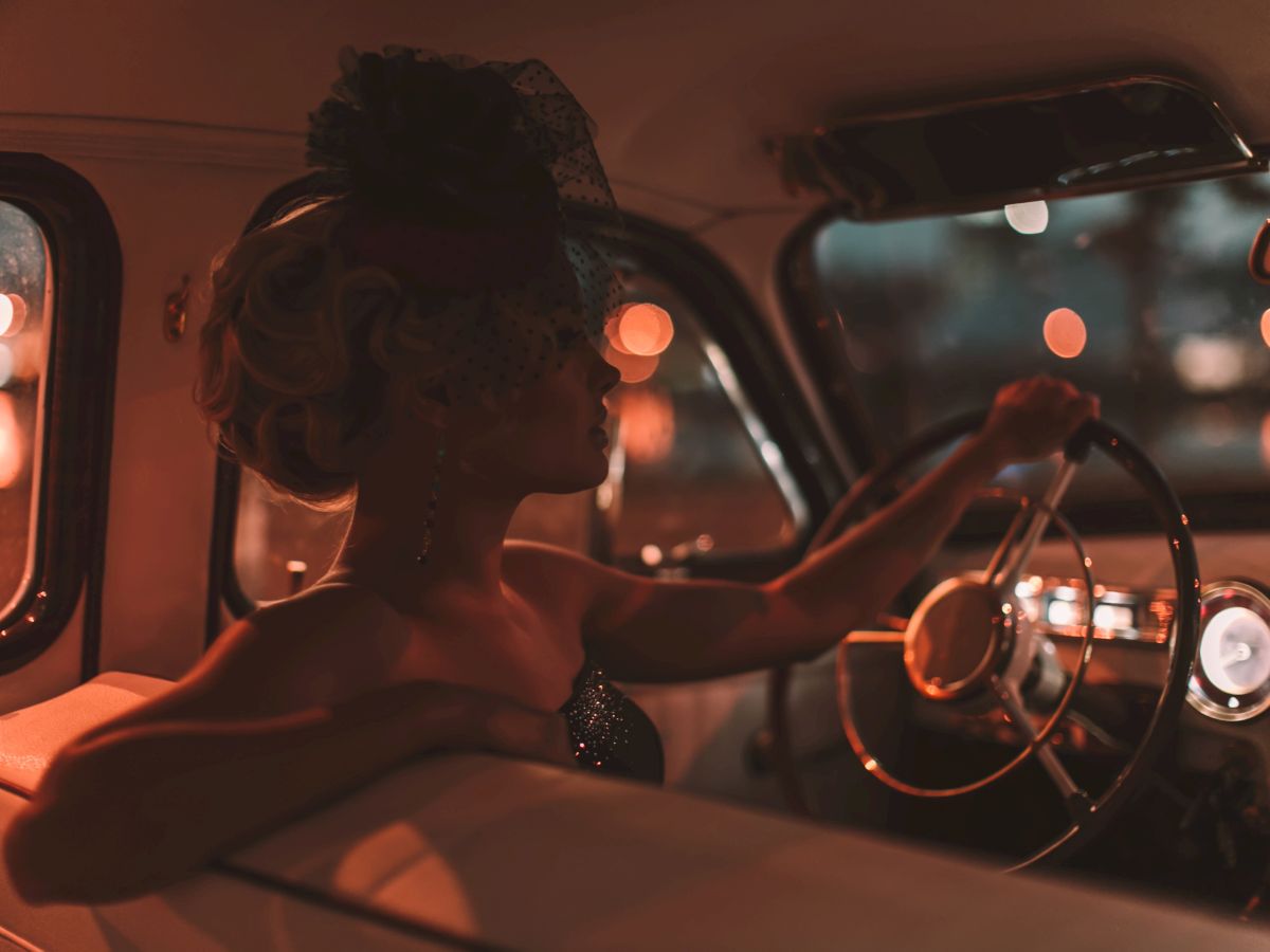 A woman in vintage attire and a hat is sitting in a classic car at night, with a cityscape blurred in the background.