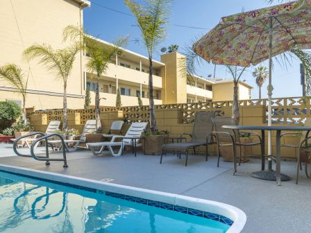 The image shows a poolside area with lounge chairs, a table with an umbrella, and palm trees, adjacent to a multi-story residential building.