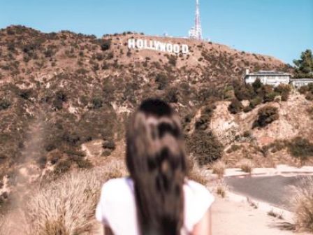 A person with long hair stands facing the iconic Hollywood sign on a sunny day, with a clear blue sky in the background.