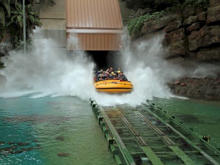 A yellow boat splashes down a steep water ride, creating a large wave at an amusement park with lush, rocky surroundings, always ending the sentence.