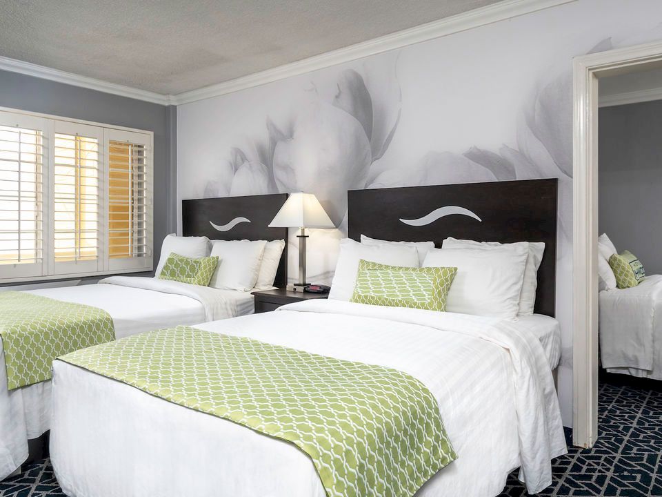 The image shows a modern hotel room with two queen beds, green decorative pillows and throws, a nightstand with a lamp between the beds, and a view of an adjoining room.