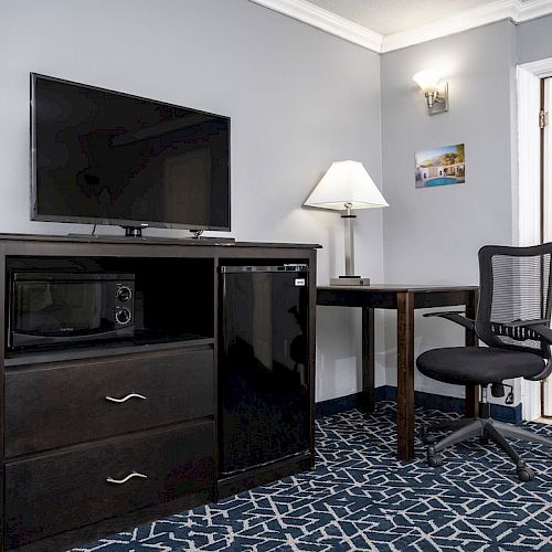 A hotel room with a TV, dresser, mini-fridge, microwave, desk with a chair, and an open door to a bathroom. The carpet has a blue pattern.