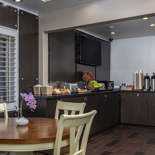 The image shows a small breakfast nook or dining area with a wooden table, chairs, counter with breakfast items, and a television on the wall.