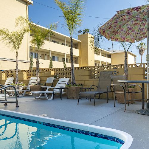 A small outdoor swimming pool with lounge chairs, tables, and an umbrella in a courtyard surrounded by a tan building and palm trees.