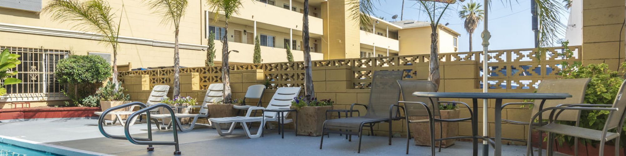 A poolside setting with lounge chairs, table with an umbrella, and palm trees against a building backdrop. Bright and sunny outdoor scene.