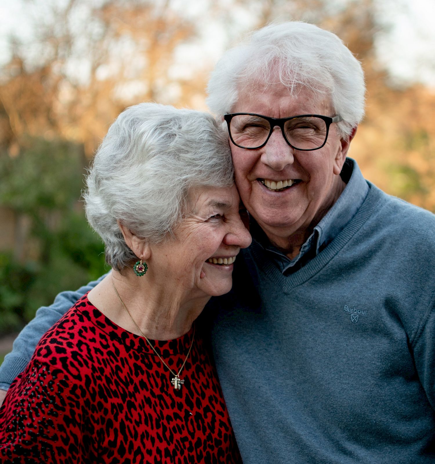 An elderly couple is happily embracing outdoors, with the woman wearing a red sweater and the man in a blue sweater.