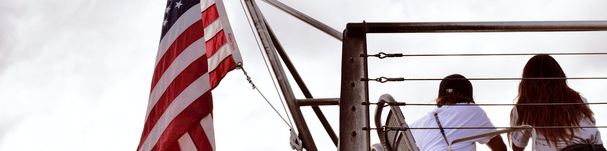 An American flag waves beside two people sitting on elevated seats, looking into the distance as clouds cover the sky.