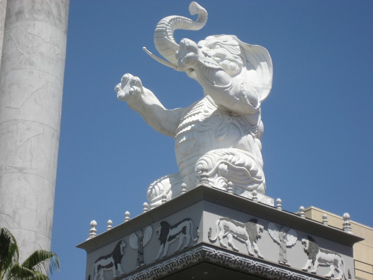 The image shows a white sculpted elephant standing on a pedestal with detailed engravings of animals, positioned against a clear blue sky.