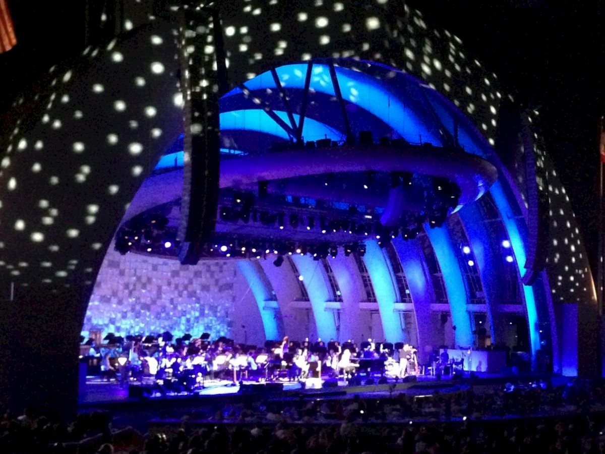 A brightly lit concert stage with musicians performing under an arched structure illuminated with blue and white lights.