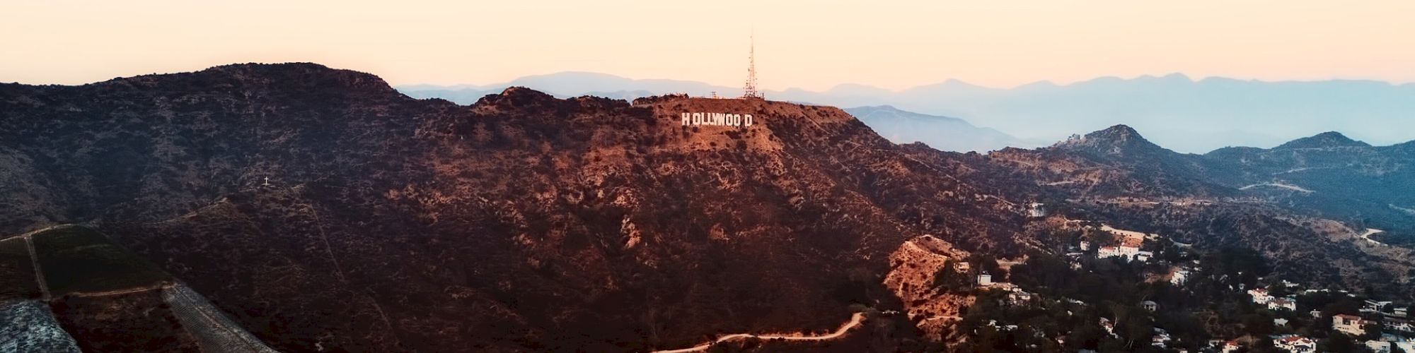 Aerial view of mountainous terrain with scattered houses and a famous landmark sign on the hilltop, during sunset with a hazy sky in the background.