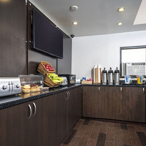 The image shows a breakfast buffet area with bagels, pastries, fruit, cereal dispensers, coffee dispensers, and a toaster on a dark countertop.