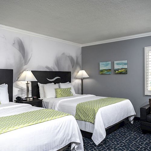 A hotel room with two beds, white linens, green accents, a black couch, wall art, lamps, and a window with blinds, on a patterned blue carpet.