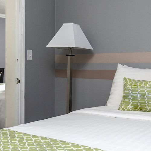 A modern hotel room with a neatly made bed featuring green-patterned pillows. A second bed and a TV are visible through the open doorway.