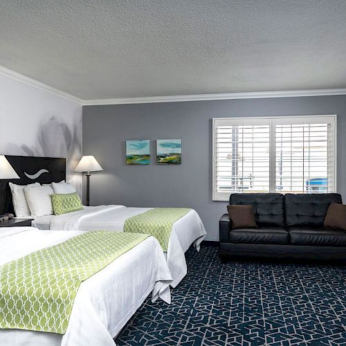 A modern hotel room with two beds with green accents, a sofa, lamps, wall art, and windows with blinds in a clean, neutral-toned setting.