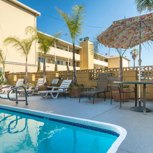 The image shows a poolside area with lounge chairs, a table with an umbrella, palm trees, and a building in the background.