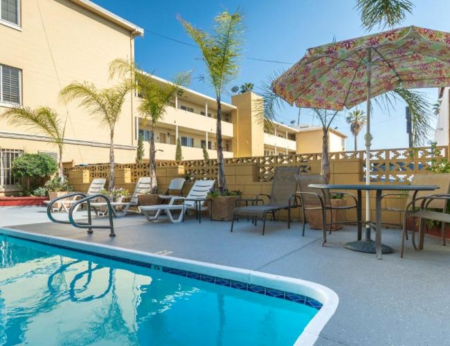 The image shows a poolside area with lounge chairs, a table with an umbrella, palm trees, and a building in the background.