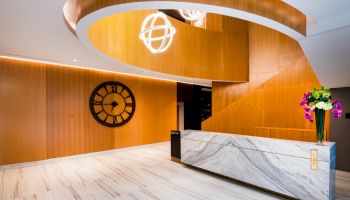 The image shows a modern reception area with wooden and marble design elements, a large wall clock, and floral arrangements on the desk.