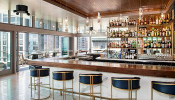 A modern bar with a wooden counter, blue and gold bar stools, an extensive liquor display, and large windows overlooking a cityscape, ending the sentence.