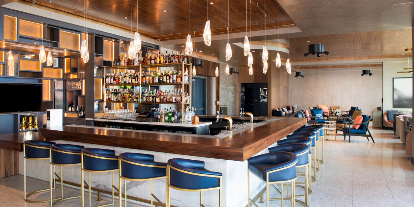 A modern bar with blue barstools, a well-stocked liquor shelf, pendant lights, and adjacent seating areas, creating a chic and inviting ambiance.