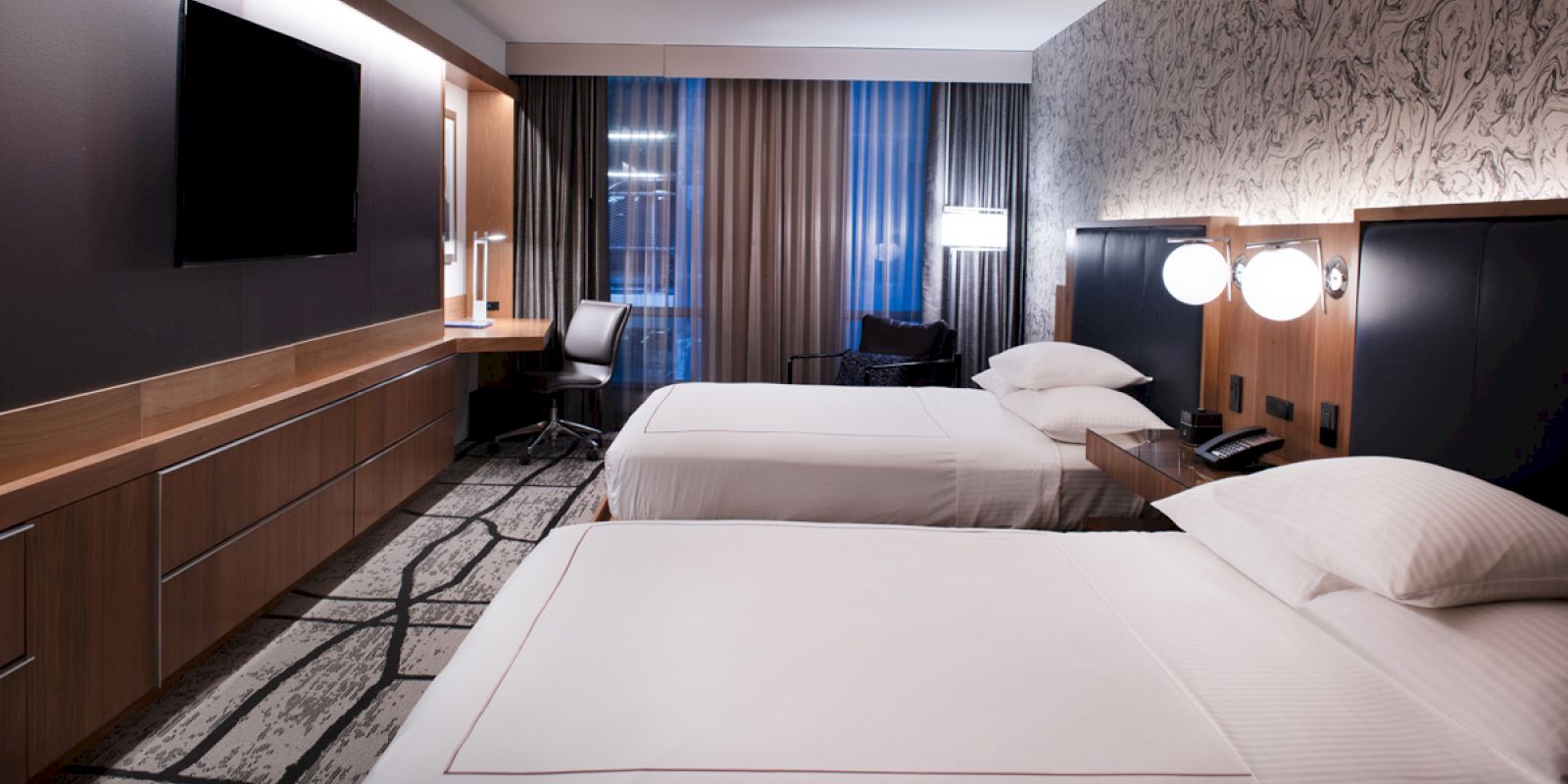 A modern hotel room with two neatly made beds, a large flat-screen TV, a desk with a chair, and contemporary decor and lighting.
