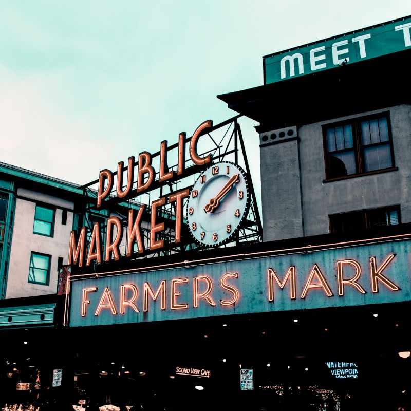 The image shows the iconic neon signs of a public market and a farmers market, with surrounding buildings and a clock displaying the time.