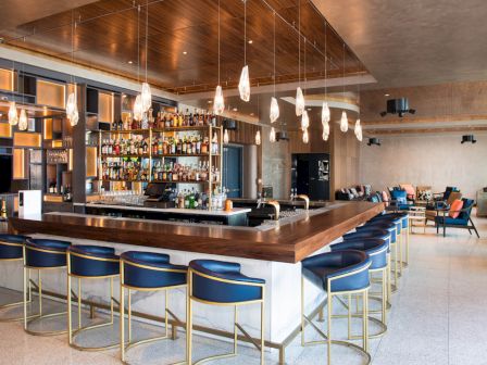This image shows a modern bar with a wooden countertop, blue barstools, and a well-stocked liquor display, lit by stylish hanging lights.