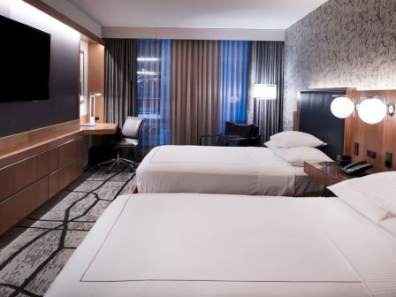 The image shows a modern hotel room with two beds, a large TV, a desk with a chair, and stylish lighting and decor, featuring a wall-mounted TV.