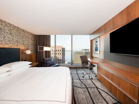 A modern hotel room with a large bed, wall-mounted TV, desk, chair, and floor-to-ceiling windows offering a city view.